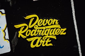 A yellow sticker with stylized, cursive text displaying 'Devon Rodriguez Art' against a dark background with white highlights.