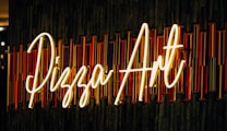Neon sign displaying the words 'Pizza Art' styled in a cursive font against a textured background with vertical colored lines. The sign is illuminated, casting a warm glow.