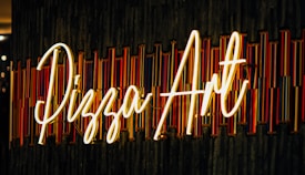 Neon sign displaying the words 'Pizza Art' styled in a cursive font against a textured background with vertical colored lines. The sign is illuminated, casting a warm glow.
