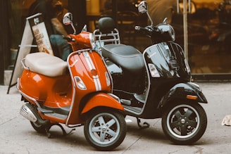 Two scooters are parked closely together on a sidewalk. One scooter is vibrant red with a beige seat, while the other is glossy black. They are positioned in front of a glass storefront with reflections visible in the windows.