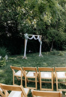 A wooden arch adorned with white drapery and floral decorations is set up on a grassy area surrounded by lush, green trees. In front of the arch, several rows of wooden chairs are neatly arranged, suggesting preparation for an outdoor ceremony.