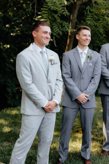 Two men in formal suits stand outside, surrounded by green foliage. Both appear to be smiling and are dressed in light-colored suits, one in a pale grey and the other in a slightly darker shade, with light-colored ties and boutonnieres.
