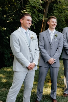 Two men in formal suits stand outside, surrounded by green foliage. Both appear to be smiling and are dressed in light-colored suits, one in a pale grey and the other in a slightly darker shade, with light-colored ties and boutonnieres.