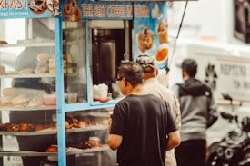 A street food cart offers a variety of baked goods and coffee to a small group of people. The cart has a blue frame and displays doughnuts and pastries behind a glass case. Three individuals are gathered around, with one person examining the menu or items closely.