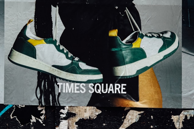 A poster depicting a pair of stylish sneakers with green, white, and yellow accents. The sneakers are set against a background featuring a person's leg and long hair. The words 'Times Square' are prominently displayed across the image.