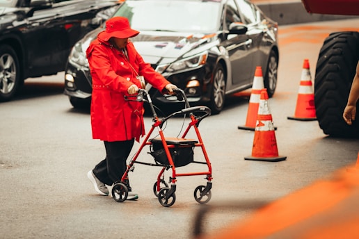 A person wearing a red coat and hat is using a red walker to navigate on a city street. Surrounding the individual are several traffic cones and moving vehicles. The scene suggests urban traffic and mobility assistance.
