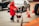 A person wearing a red coat and hat is using a red walker to navigate on a city street. Surrounding the individual are several traffic cones and moving vehicles. The scene suggests urban traffic and mobility assistance.
