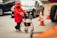 A person wearing a red coat and hat is using a red walker to navigate on a city street. Surrounding the individual are several traffic cones and moving vehicles. The scene suggests urban traffic and mobility assistance.