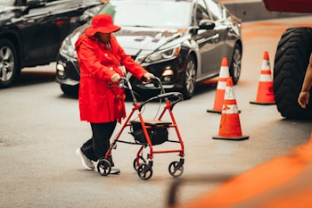 A person wearing a red coat and hat is using a red walker to navigate on a city street. Surrounding the individual are several traffic cones and moving vehicles. The scene suggests urban traffic and mobility assistance.
