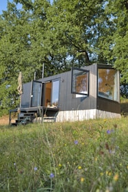 A modern tiny house with large windows is situated amidst a lush green landscape. The structure appears to be a compact living space, featuring an outdoor seating area with a patio umbrella. It is surrounded by tall trees and a grassy field dotted with small wildflowers.