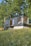 A modern tiny house with large windows is situated amidst a lush green landscape. The structure appears to be a compact living space, featuring an outdoor seating area with a patio umbrella. It is surrounded by tall trees and a grassy field dotted with small wildflowers.