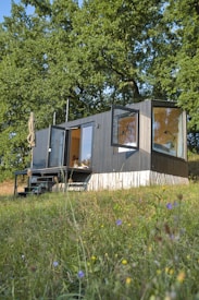 A modern tiny house with large windows is situated amidst a lush green landscape. The structure appears to be a compact living space, featuring an outdoor seating area with a patio umbrella. It is surrounded by tall trees and a grassy field dotted with small wildflowers.