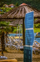 A scenic lakeside setting with several lounge chairs arranged on a sandy area. Straw umbrellas provide shade, and a palm tree adds a tropical touch. In the background, a body of water is visible with small boats docked and lush greenery surrounding the area. A blue sign with blurred text is in the foreground.