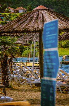 A scenic lakeside setting with several lounge chairs arranged on a sandy area. Straw umbrellas provide shade, and a palm tree adds a tropical touch. In the background, a body of water is visible with small boats docked and lush greenery surrounding the area. A blue sign with blurred text is in the foreground.