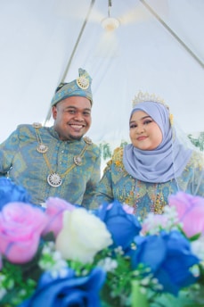 A couple dressed in traditional formal attire smiles at the camera. The man is wearing a hat with an ornate design, while the woman wears a crown and a hijab with intricate patterns. In the foreground, there are vibrant, colorful flowers, including blue and pink roses.