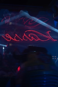 Neon red light forms abstract shapes in a dark environment, with soft blue lighting providing contrast. A hint of machinery or industrial equipment is visible in the lower part of the image.