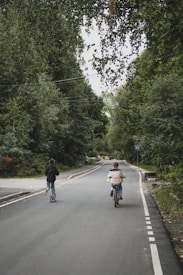 A quiet road surrounded by lush green trees. Two people are traveling down the road; one is riding a bicycle, and the other is using a scooter. The scene exudes a serene and tranquil atmosphere, with various shades of green from the surrounding forest.