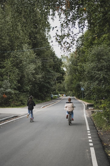 A quiet road surrounded by lush green trees. Two people are traveling down the road; one is riding a bicycle, and the other is using a scooter. The scene exudes a serene and tranquil atmosphere, with various shades of green from the surrounding forest.