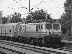 A monochrome image showing a railway engine attached to a series of passenger compartments on train tracks. The engine has multiple headlights and what seems to be a stylized text written in a non-English script. There is visible overhead wiring for electric trains, with trees and some structures in the background.