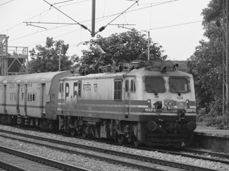 A monochrome image showing a railway engine attached to a series of passenger compartments on train tracks. The engine has multiple headlights and what seems to be a stylized text written in a non-English script. There is visible overhead wiring for electric trains, with trees and some structures in the background.