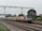 A locomotive with several passenger cars travels along a set of railway tracks. Overhead electrical lines and a metal gantry are visible above the train. Lush green trees line the tracks, and a station sign reads 'Dhandera' in multiple scripts. The sky is overcast, adding a sense of calm to the scene.