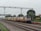 A locomotive with several passenger cars travels along a set of railway tracks. Overhead electrical lines and a metal gantry are visible above the train. Lush green trees line the tracks, and a station sign reads 'Dhandera' in multiple scripts. The sky is overcast, adding a sense of calm to the scene.