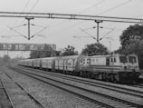 A long train is traveling on a set of railway tracks, with visible overhead power lines and a supportive metal structure. The scene includes trees lining the tracks, while the train moves through an open, rural setting. The train itself is marked with text in a non-English script.