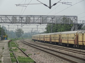 A long, beige-colored passenger train with red and maroon accents is moving along railway tracks. Overhead, there are electrical wires and a metal pedestrian overpass. The tracks are surrounded by greenery and trees, with a small platform and a yellow signboard on the left side of the image. The weather appears to be overcast.