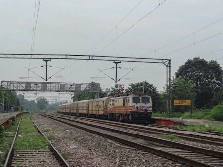 A long train with multiple carriages travels along a railway track surrounded by greenery and trees. Overhead electrical wires and gantries are visible, indicating an electrified railway. The sky is overcast, and a yellow station sign reads 'Dhandhera' in both English and a regional script.