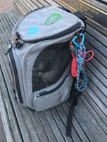 A gray backpack with embroidered patches is resting on a wooden bench. The bag has multiple compartments, one of which is mesh, and a carabiner attached to colorful climbing ropes hanging from it.