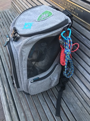 A gray backpack with embroidered patches is resting on a wooden bench. The bag has multiple compartments, one of which is mesh, and a carabiner attached to colorful climbing ropes hanging from it.