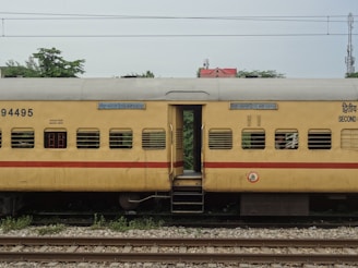 A yellow passenger train coach with a red stripe is seen stationary on railway tracks. The coach has several windows with bars and signage in multiple languages. There are some trees and a building with a red roof visible in the background.