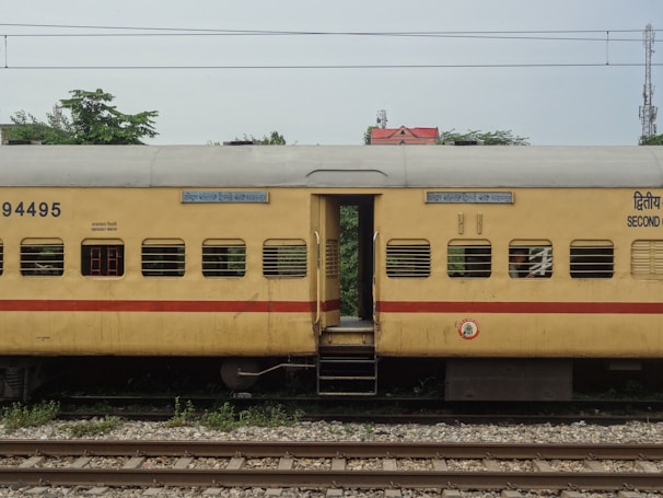 A yellow passenger train coach with a red stripe is seen stationary on railway tracks. The coach has several windows with bars and signage in multiple languages. There are some trees and a building with a red roof visible in the background.