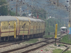 A train with yellow and brown coaches is moving along railway tracks beneath a network of overhead wires. Passengers can be seen leaning out of the windows. To the right of the tracks, there are multiple yellow signs with 'STOP' written on them. In the distance, there is greenery and a small structure with a white roof, with a few people standing near it.