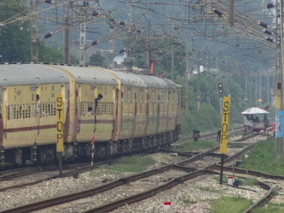 A train with yellow and brown coaches is moving along railway tracks beneath a network of overhead wires. Passengers can be seen leaning out of the windows. To the right of the tracks, there are multiple yellow signs with 'STOP' written on them. In the distance, there is greenery and a small structure with a white roof, with a few people standing near it.
