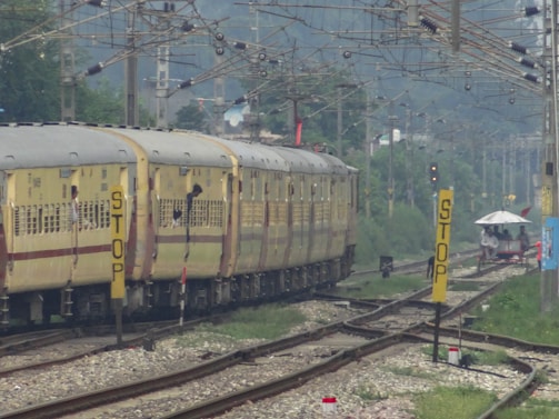 A train with yellow and brown coaches is moving along railway tracks beneath a network of overhead wires. Passengers can be seen leaning out of the windows. To the right of the tracks, there are multiple yellow signs with 'STOP' written on them. In the distance, there is greenery and a small structure with a white roof, with a few people standing near it.