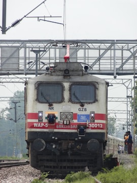 A locomotive painted in white and red with the inscription 'WAP-5 GHAZIABAD 30012' is positioned on railway tracks. Overhead electrical cables and structures are visible, providing power to the train. In the background, there is a train coach and a group of people standing near the tracks. The scene is outdoors, surrounded by greenery and a metal footbridge overhead.