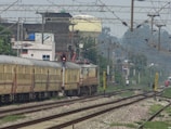 A train moves along a set of railway tracks, passing through an urban area. Overhead wires and electrical poles are visible above the train. In the background, residential and industrial buildings line the tracks. There are signals and signage near the tracks, with a few people standing by the railway lines.