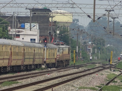 A train moves along a set of railway tracks, passing through an urban area. Overhead wires and electrical poles are visible above the train. In the background, residential and industrial buildings line the tracks. There are signals and signage near the tracks, with a few people standing by the railway lines.