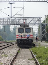 A train is positioned on railway tracks under an overhead power line structure. The locomotive has a white body with red stripes and is labeled WAP-5. Surrounding the train, there is vegetation and gravel alongside the tracks. A person is visible in the distance on the side of the tracks.