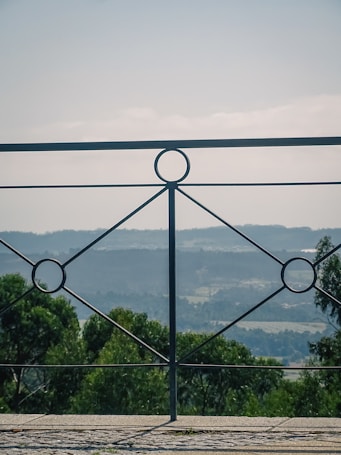 A wrought iron railing with a geometric design is in the foreground, set against a backdrop of rolling hills and a forested landscape. The sky is clear with a soft, diffused light, adding a serene atmosphere to the scene.