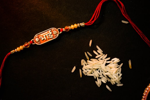 A decorative bracelet with a red thread and intricate beads, featuring an ornamental piece with text in Hindi, placed on a dark surface next to scattered grains of rice.