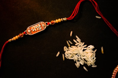 A decorative bracelet with a red thread and intricate beads, featuring an ornamental piece with text in Hindi, placed on a dark surface next to scattered grains of rice.