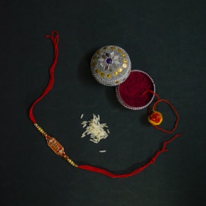 An ornate box with decorative patterns containing red vermillion powder is placed next to a red and gold thread with beads, likely a rakhi. A small pile of rice grains is also visible nearby, possibly for a traditional ceremony.