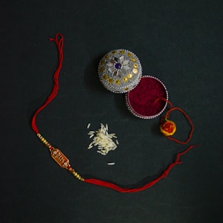 An ornate box with decorative patterns containing red vermillion powder is placed next to a red and gold thread with beads, likely a rakhi. A small pile of rice grains is also visible nearby, possibly for a traditional ceremony.