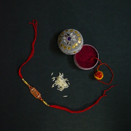 An ornate box with decorative patterns containing red vermillion powder is placed next to a red and gold thread with beads, likely a rakhi. A small pile of rice grains is also visible nearby, possibly for a traditional ceremony.