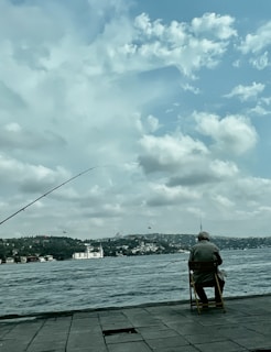 An elderly person sits on a chair by a waterfront, holding a fishing rod. The scene includes a body of water with a cityscape in the background under a sky filled with fluffy clouds. A fishing line is cast into the water, and there are buildings on a distant hill across the water.