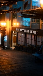 A warmly lit street scene at night shows the entrance of The Pottinger Hotel, with a glowing streetlamp and hanging lanterns casting a soft glow. The surrounding architecture features large windows reflecting ambient light, and the cobblestone path leads to the hotel entrance.