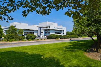 A modern college building is surrounded by lush greenery, including well-maintained lawns and trees. The structure features a sleek, white facade with large windows. Several cars are parked nearby, and a clear blue sky enhances the setting.