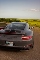 A sleek grey Porsche 911 Turbo is parked on the side of a road, with a scenic countryside landscape in the background featuring green fields and sparse trees. The license plate indicates it is from Puerto Rico.